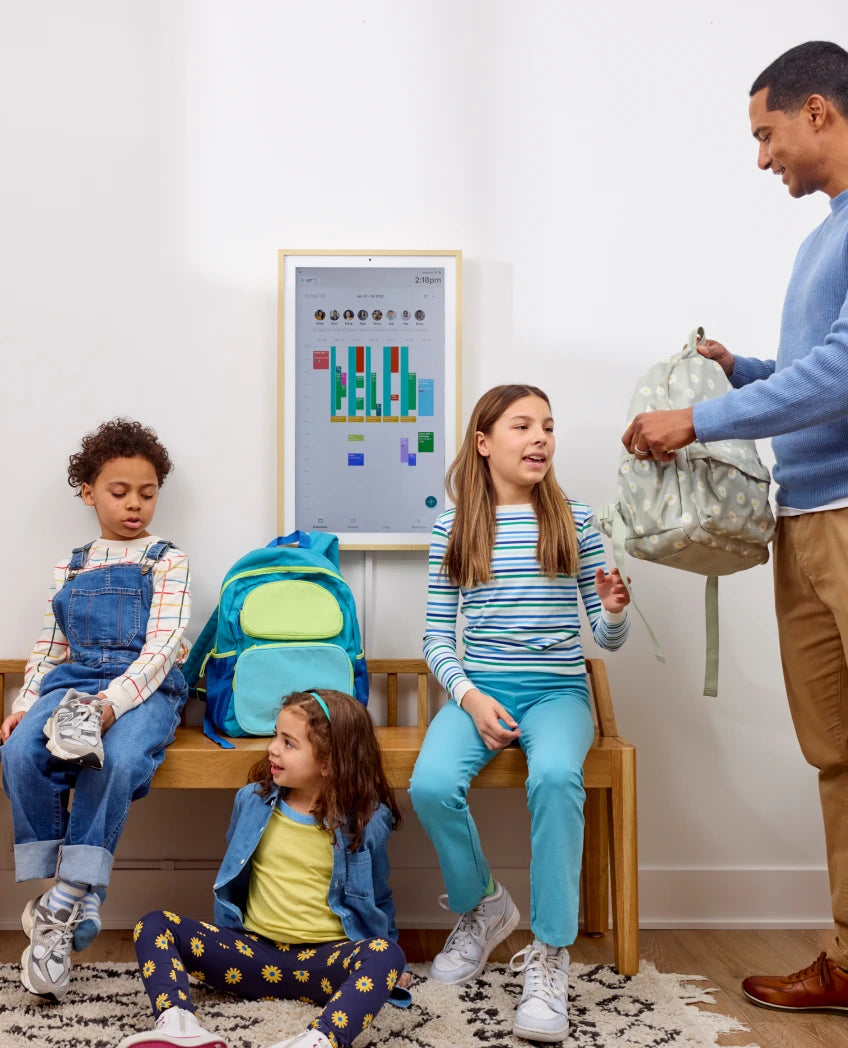 Family with children sitting on a bench with backpacks, a man standing nearby.